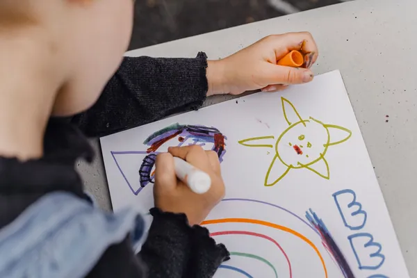 A child's hands coloring a sun and rainbow with markers