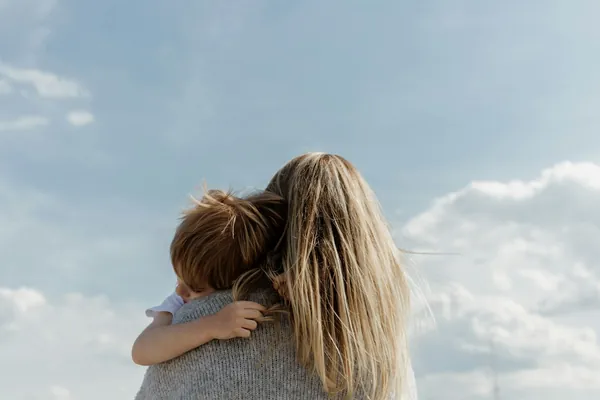 A child hugging a parent from behind, against a cloudy sky