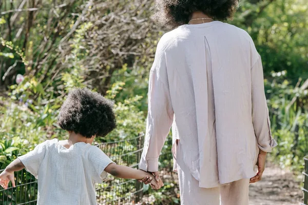 A parent and small child walking hand in hand down a sunny path, seen from behind