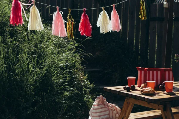 A backyard picnic table set for a party with paper tassels strung above it